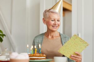 Frau sitzt am Tisch mit Geburtstagstorte, liest eine Geburtstagskarte und lächelt