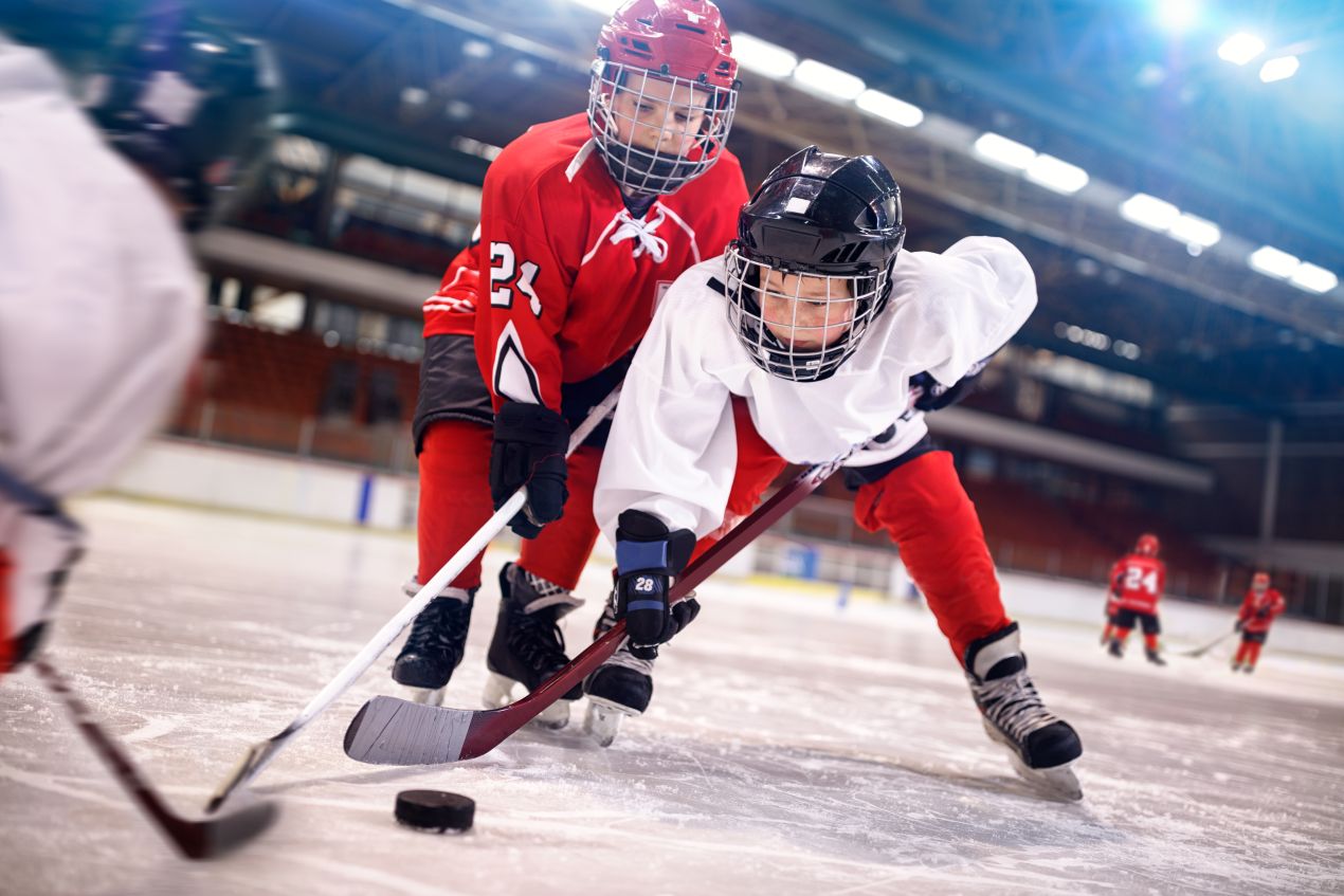 Zwei junge Eishockeyspieler duellieren sich um den Puck auf dem Spielfeld, waehrend andere Kinder im Hintergrund trainieren.